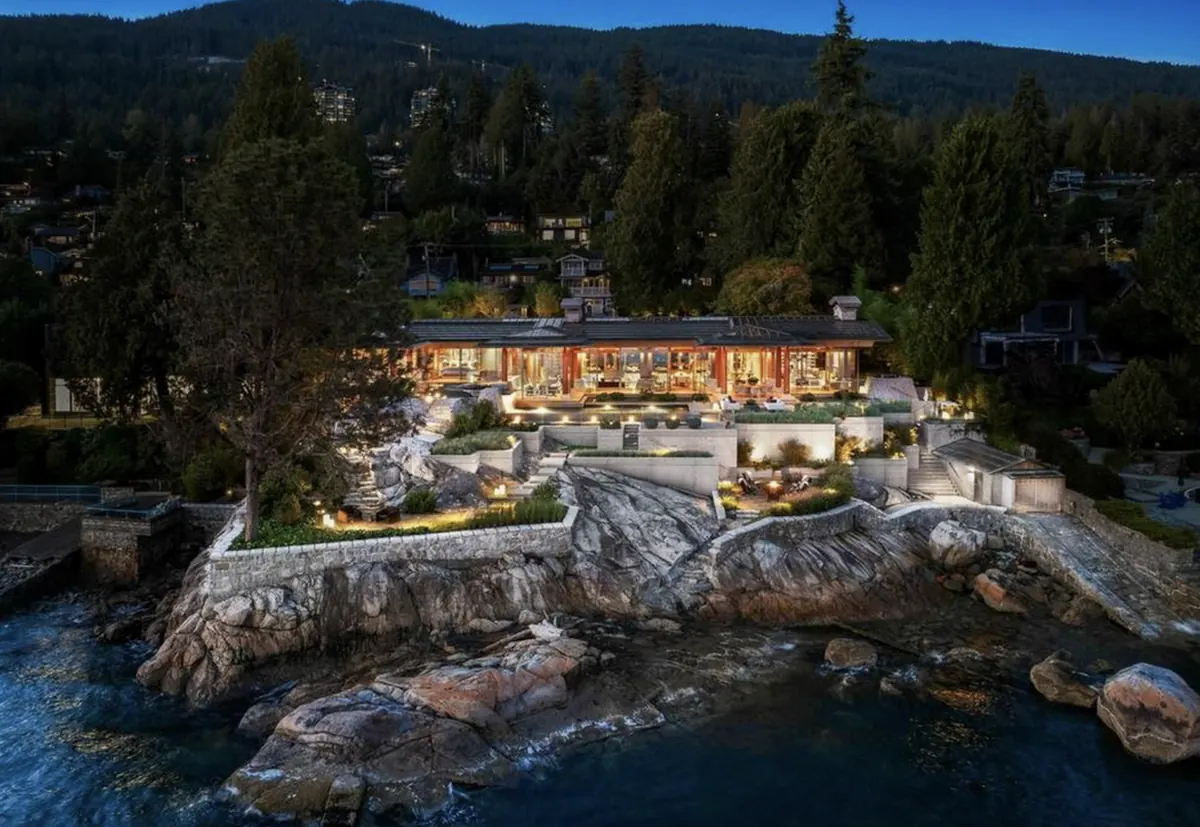 A low modern house lit at dusk on a rocky waterfront point, seen from the water
