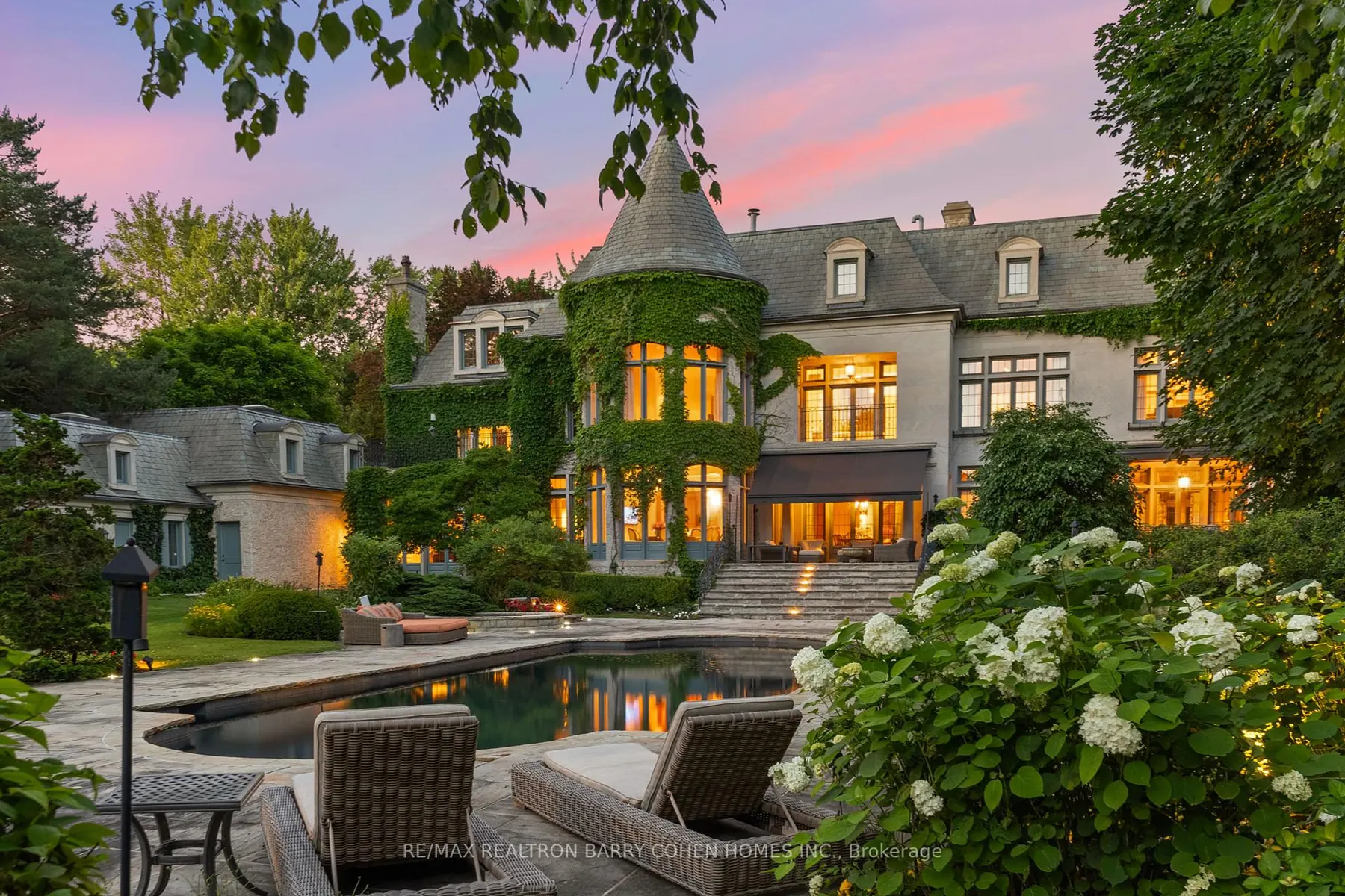 A turreted stone mansion lit at dusk, with a swimming pool and hydrangeas in the foreground