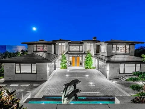 A large contemporary house floodlit at night, with a lit driveway, pool and sculpture