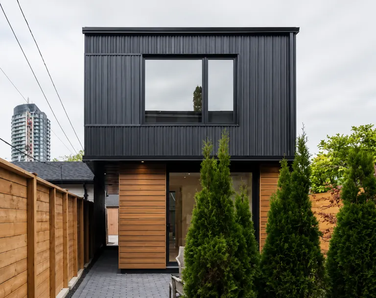 Finished two-storey laneway suite seen from the garden, a black metal-clad upper floor with a wide window over a ground floor in cedar and full-height glazing