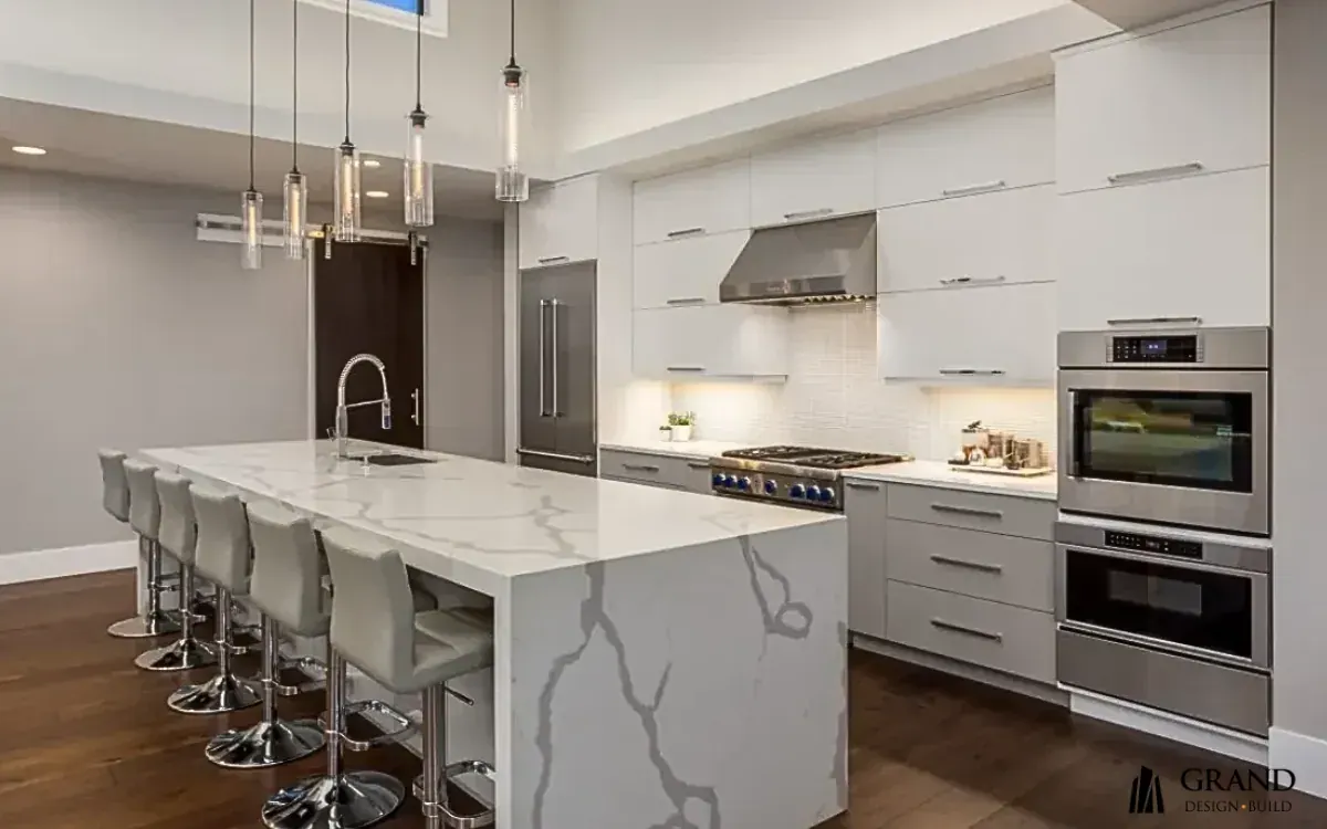 A finished Grand Design Build kitchen with a long marble waterfall island, upholstered stools, linear glass pendants, handleless white cabinets and stainless wall ovens